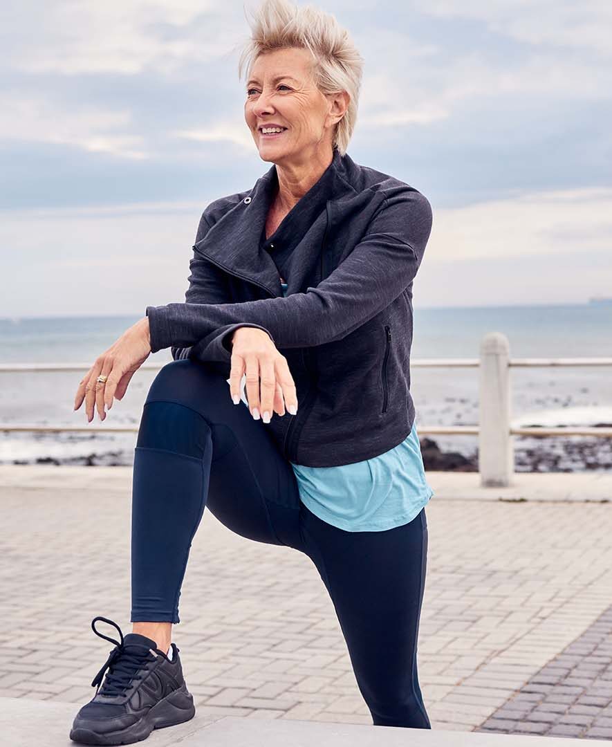 Eine ältere, aktive Frau mit kurzem, blondem Haar trägt sportliche Kleidung und dehnt sich am Meer. Sie lehnt sich entspannt auf ihr Knie, während sie lächelt und in die Ferne blickt. Die Szene strahlt Vitalität, Lebensfreude und Wohlbefinden aus. Im Hintergrund erstreckt sich das Meer unter einem bewölkten Himmel.